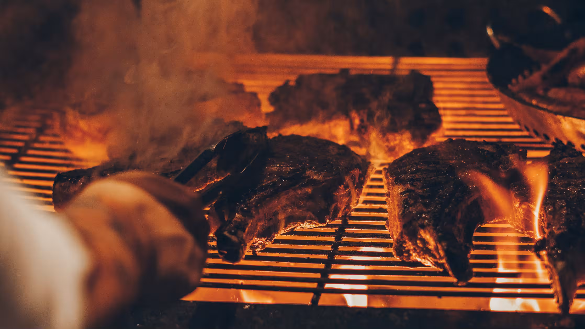 Steaks over the open grill, flames through the bars, a cook turning one with tongs.